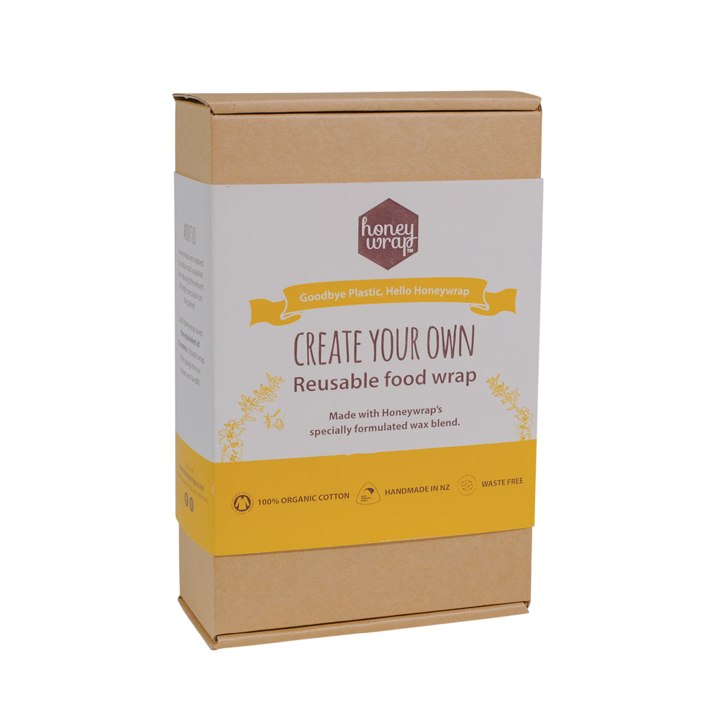 A kit labeled 'Create Your Own Reusable Food Wrap' by Honeywrap, consisting of a wax block, mini grater, cotton squares, printed fabric, felt tip pens, and a paintbrush, all packaged in a brown cardboard box.