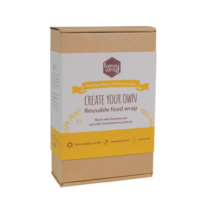 A kit labeled 'Create Your Own Reusable Food Wrap' by Honeywrap, consisting of a wax block, mini grater, cotton squares, printed fabric, felt tip pens, and a paintbrush, all packaged in a brown cardboard box.