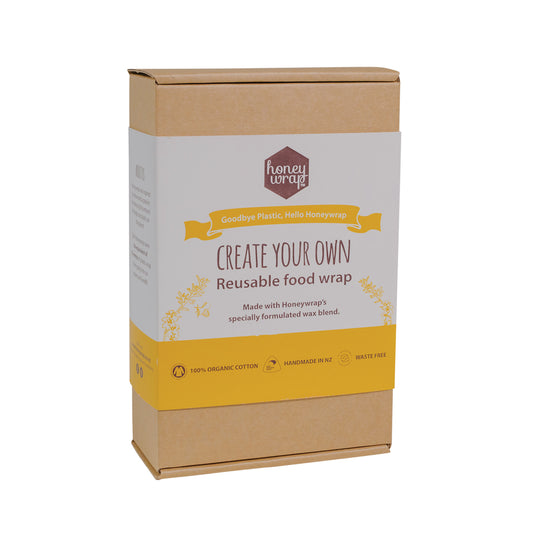 A kit labeled 'Create Your Own Reusable Food Wrap' by Honeywrap, consisting of a wax block, mini grater, cotton squares, printed fabric, felt tip pens, and a paintbrush, all packaged in a brown cardboard box.